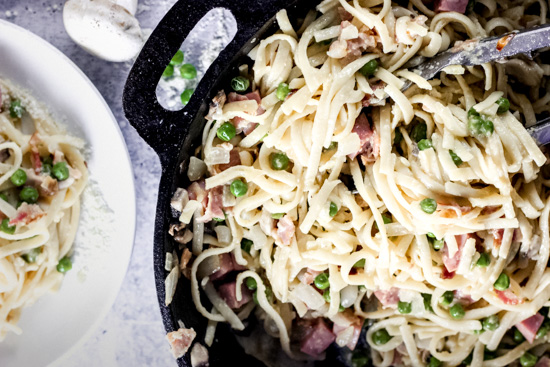 overhead view of a pan full of linguine with noodles, bacon, ham, mushrooms, peas