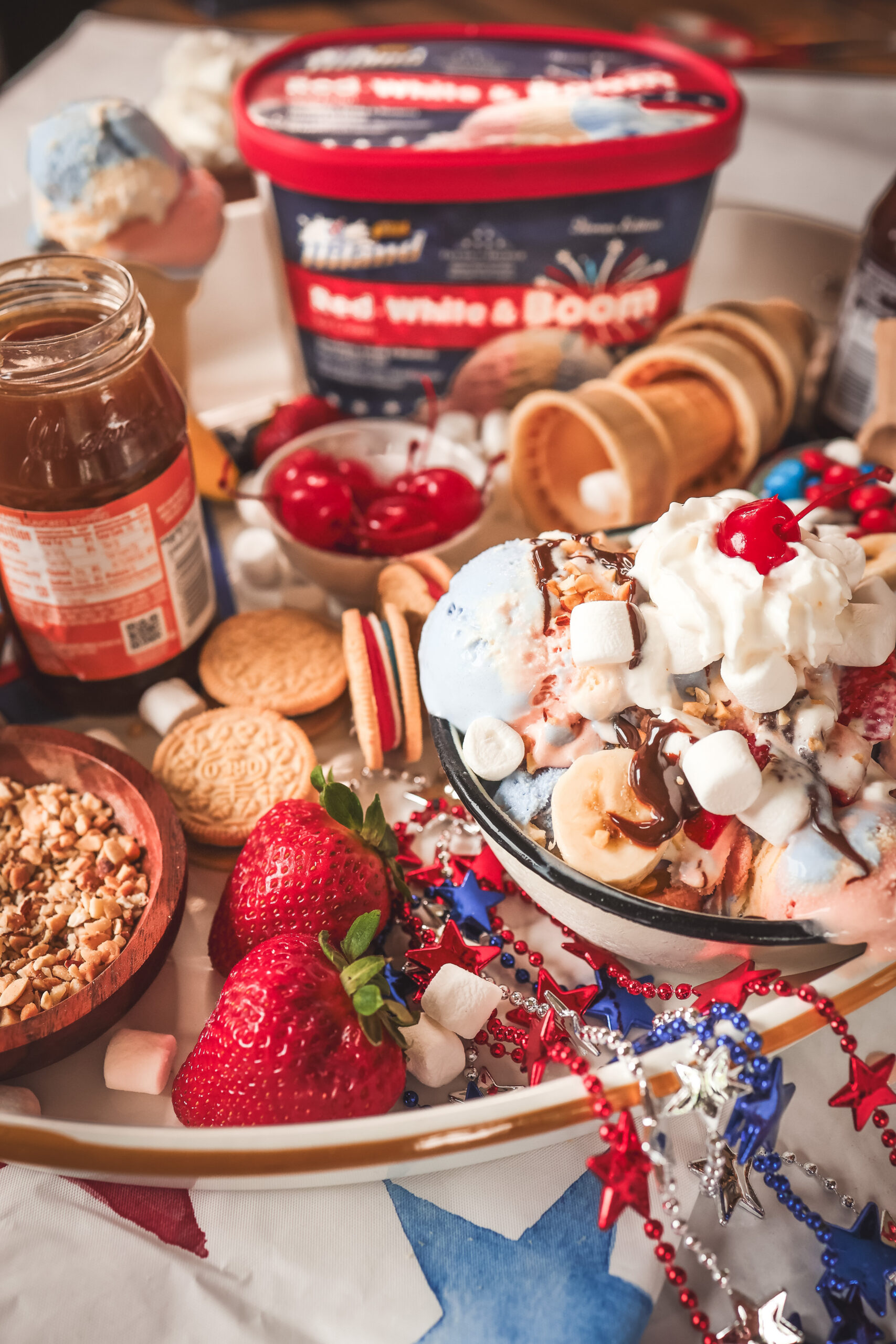 4th of July ice cream sundae board with Hiland Dairy Red White and Boom ice cream surrounded by colorful toppings including hot fudge, sprinkles, strawberries, blueberries, and whipped cream