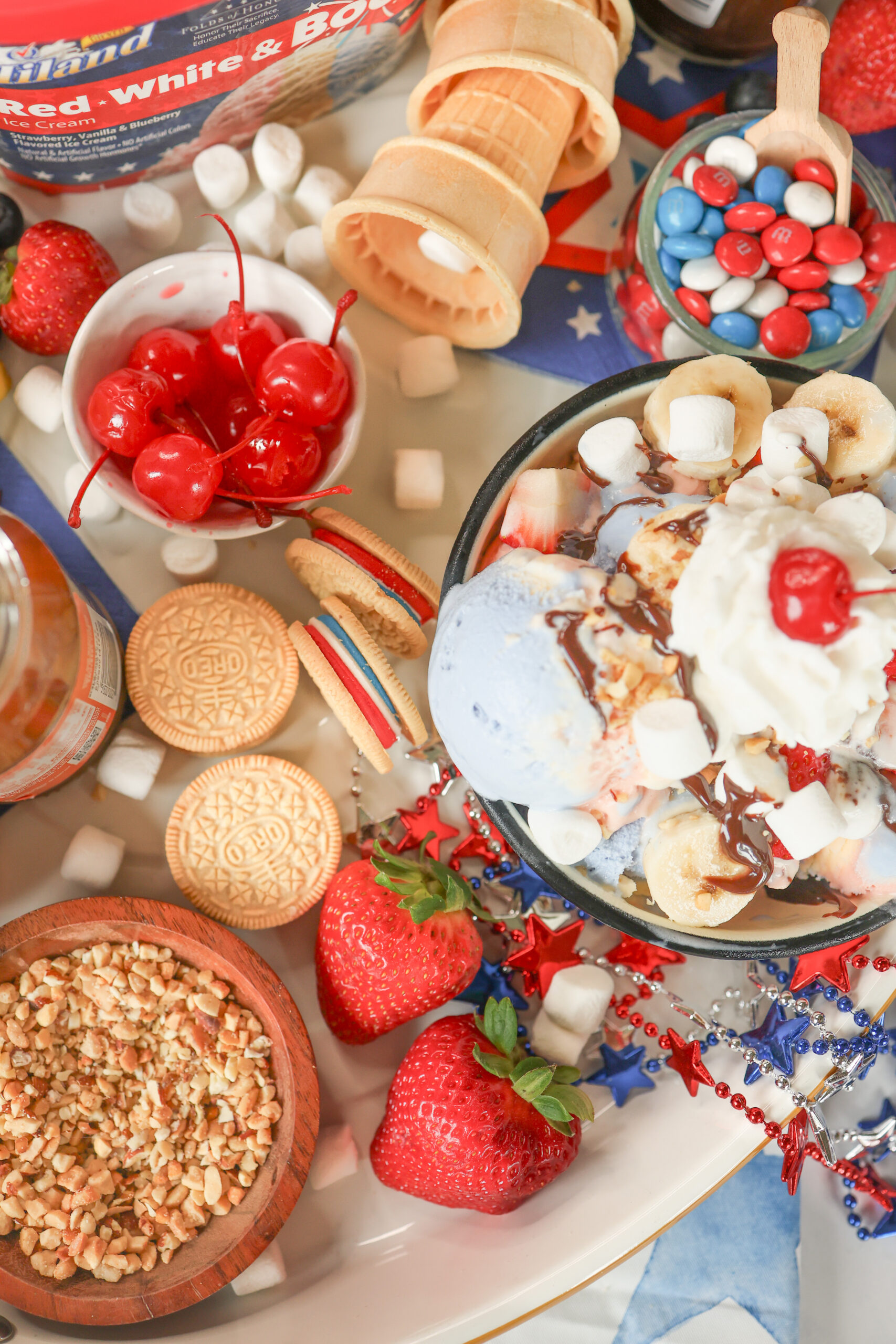4th of July ice cream sundae board with Hiland Dairy Red White and Boom ice cream surrounded by colorful toppings including hot fudge, sprinkles, strawberries, blueberries, and whipped cream