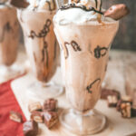 A straight on shot of 3 milkshake glasses filled with a snickers milkshake topped with chocolate drizzle and a snickers bite